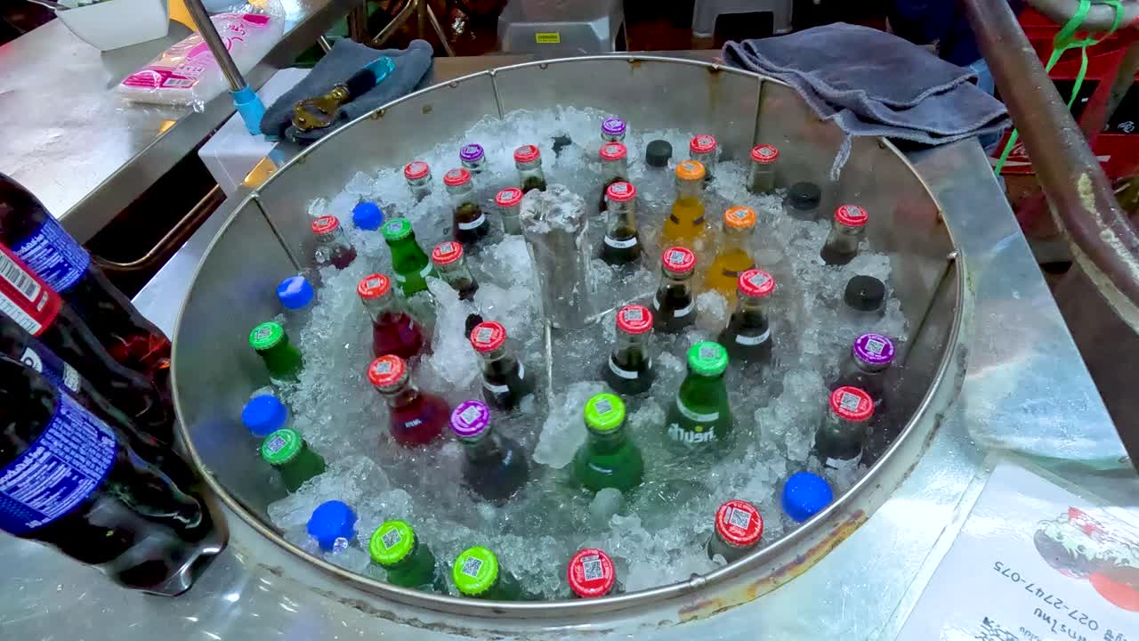 Colorful glass soft drink bottles spin in ice water under bright market lighting, overhead view
