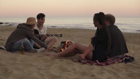 Young-cheerful-friends-sitting-by-the-fire-on-the-beach-in-the-evening,-drinking-beer-and-playing-guitar.-Shot-in-4k