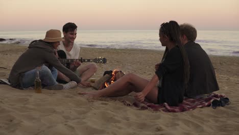 young cheerful friends sitting by the fire on the beach in the evening, drinking beer and playing guitar. shot in 4k