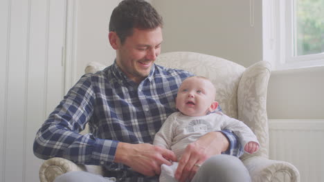 father cuddling smiling baby son as they sit in chair at home together - shot in low motion