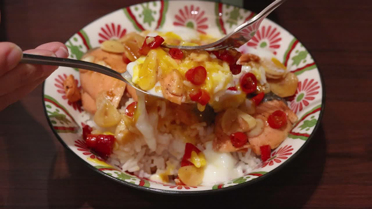 Hand uses spoon to mix cooked salmon, scrambled egg, garlic, chili, and rice in a decorative bowl under warm indoor lighting, top-down view
