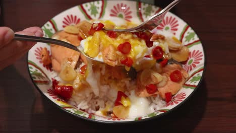 hand uses spoon to mix cooked salmon, scrambled egg, garlic, chili, and rice in a decorative bowl under warm indoor lighting, top-down view
