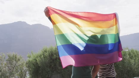 una feliz pareja masculina diversa sosteniendo una bandera lgbt con los brazos abiertos