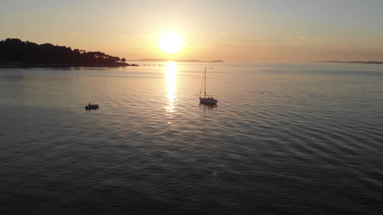 Un sole dorato proietta i suoi raggi di luce sull'acqua, illuminando uno yacht ancorato al largo della costa. Mari calmi lambiscono la riva baciata dal sole vicino alla spiaggia di Bueu, Galizia, Spagna.
