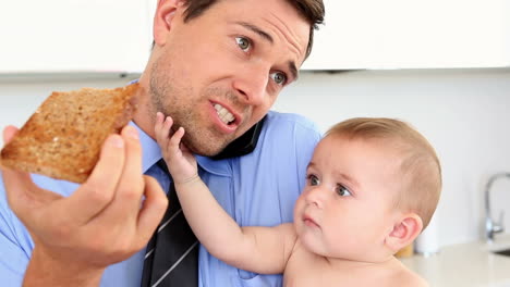 hombre de negocios sosteniendo a su bebé y hablando por teléfono mientras come tostadas