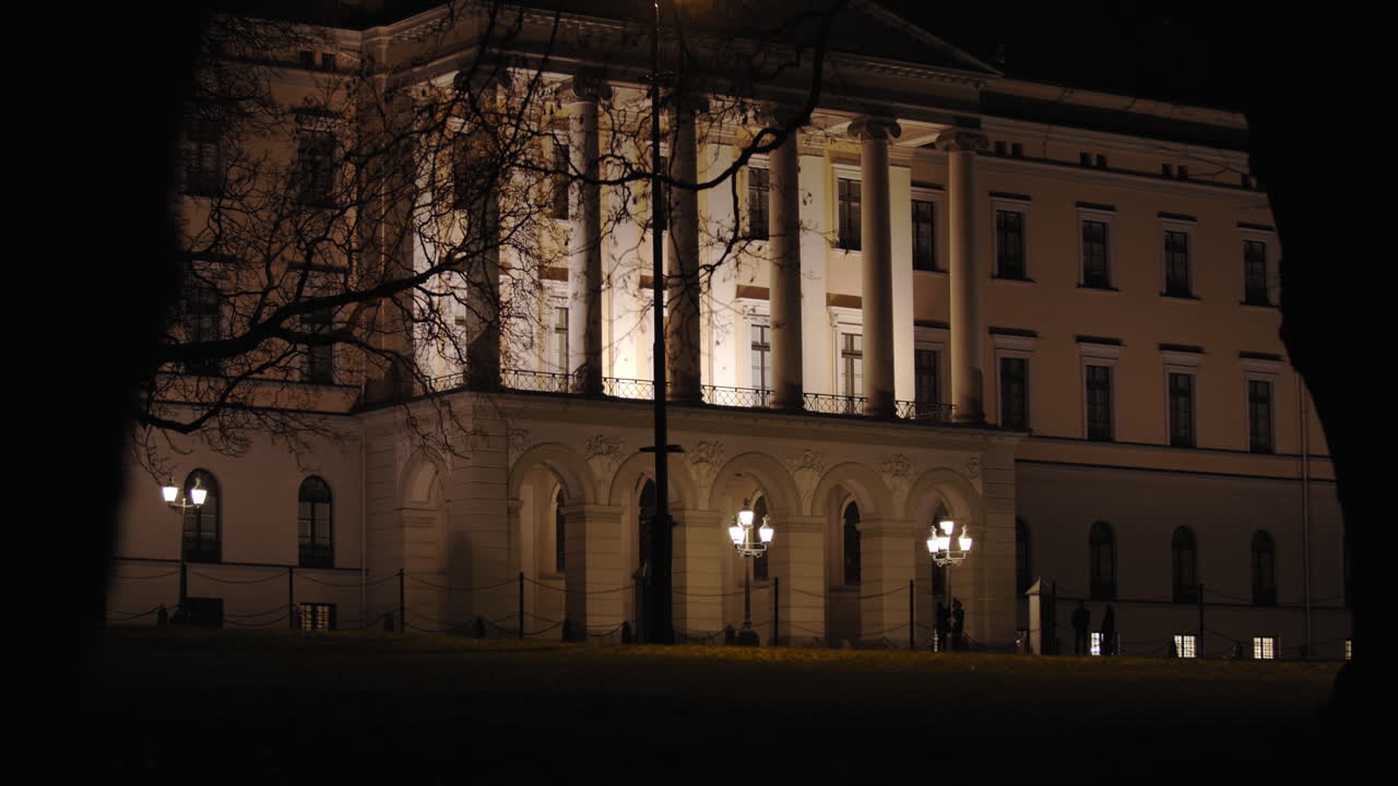 Cinematic wide to close-up 4K shot of dark trees moving in parallax motion in front of the illuminated Norwegian Royal Palace with armed guard walking in front of building, at night in Oslo Norway