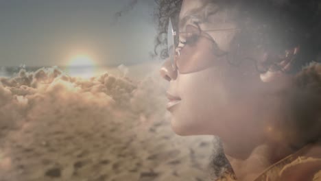 animation of african american woman wearing sunglasses at beach, over clouds