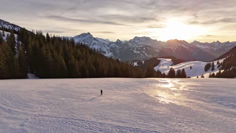 Aufsteigende-Drohnenaufnahme,-Die-Eine-Person-Zeigt,-Die-Bei-Goldenem-Sonnenuntergang-Auf-Einem-Verschneiten-Hang-In-Den-Schweizer-Bergen-Läuft