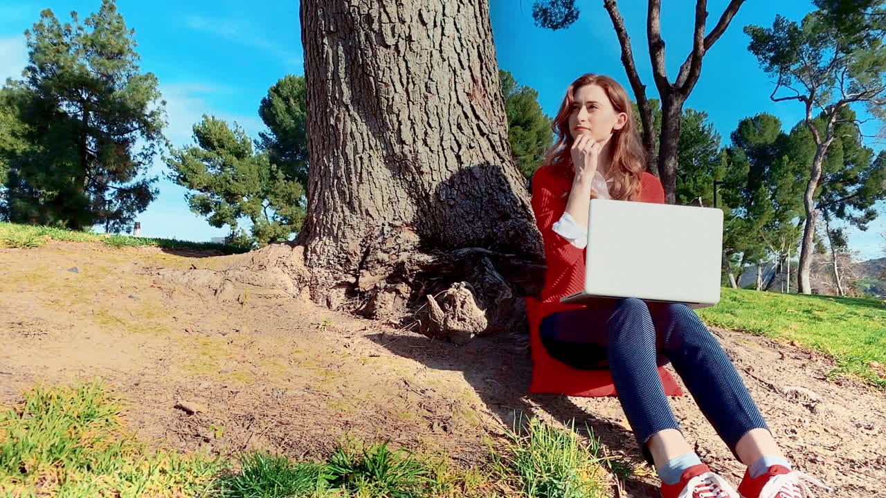 una mujer feliz, atractiva, joven y caucásica en el parque, escribiendo en una computadora portátil debajo de un árbol