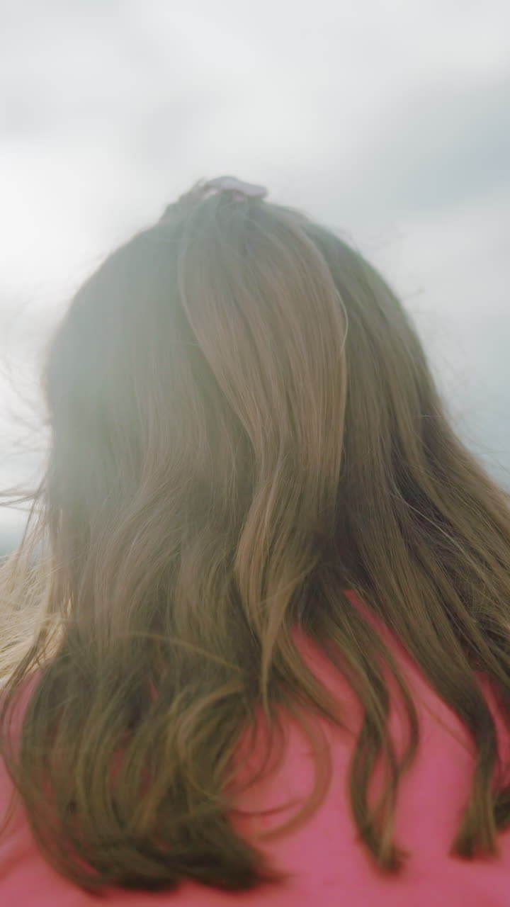 핑크색 옷을 입은 여성의 뒷모습, 긴 파란 머리카락이 아래로 떨어지고, 따뜻한 빛에 비추어, 부드러운 자연 빛은 흐릿한 나무와 열린 하늘로 평온한 시골 분위기를 만니다.