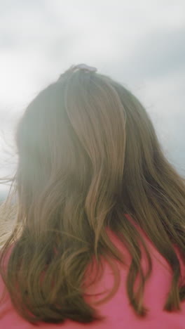 vista trasera de una mujer de color rosa con el cabello largo y ondulado en cascada hacia abajo, iluminada por la cálida luz del sol, el suave resplandor natural crea una atmósfera serena de campo con árboles borrosos y cielo abierto