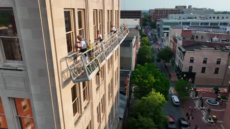 blue collar workers stand on scaffolding painting windows of an urban office building