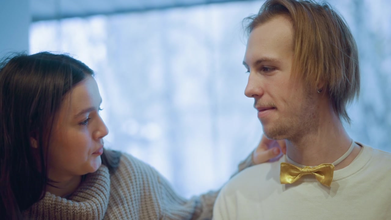 Close up of woman with long dark hair wearing knitted sweater leaning towards man adjusting his golden bow tie. Soft lighting creates warm indoor atmosphere background blurred to emphasize