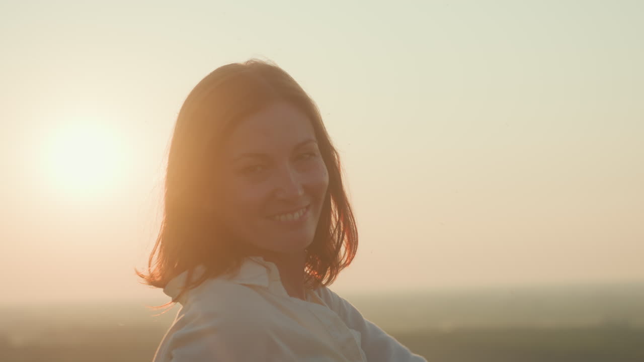 cheerful lady standing under open sky with soft sunlight glowing around hair as she turns with subtle smile, capturing serene mood in calm countryside during golden hour