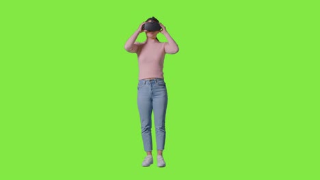 full length shot of woman putting on virtual reality headset and looking around against green screen studio background