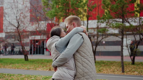 partner bonding moment between couple as man lifts woman warmly in joyful embrace, holding her close while they kiss affectionately outdoors with grass, trees, and parked cars visible in background