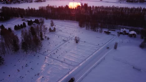 aerial view during winter in north of sweden, flying over a road with cars and fields covered in snow slowly tilting up revealing a frozen lake and the sun setting behind the mountains