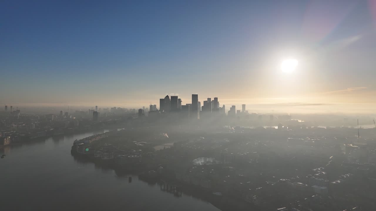 Aerial drone footage soaring over the River Thames towards Canary Wharf skyline, with misty sunlight illuminating the city. Perfect for cinematic urban stock videos.