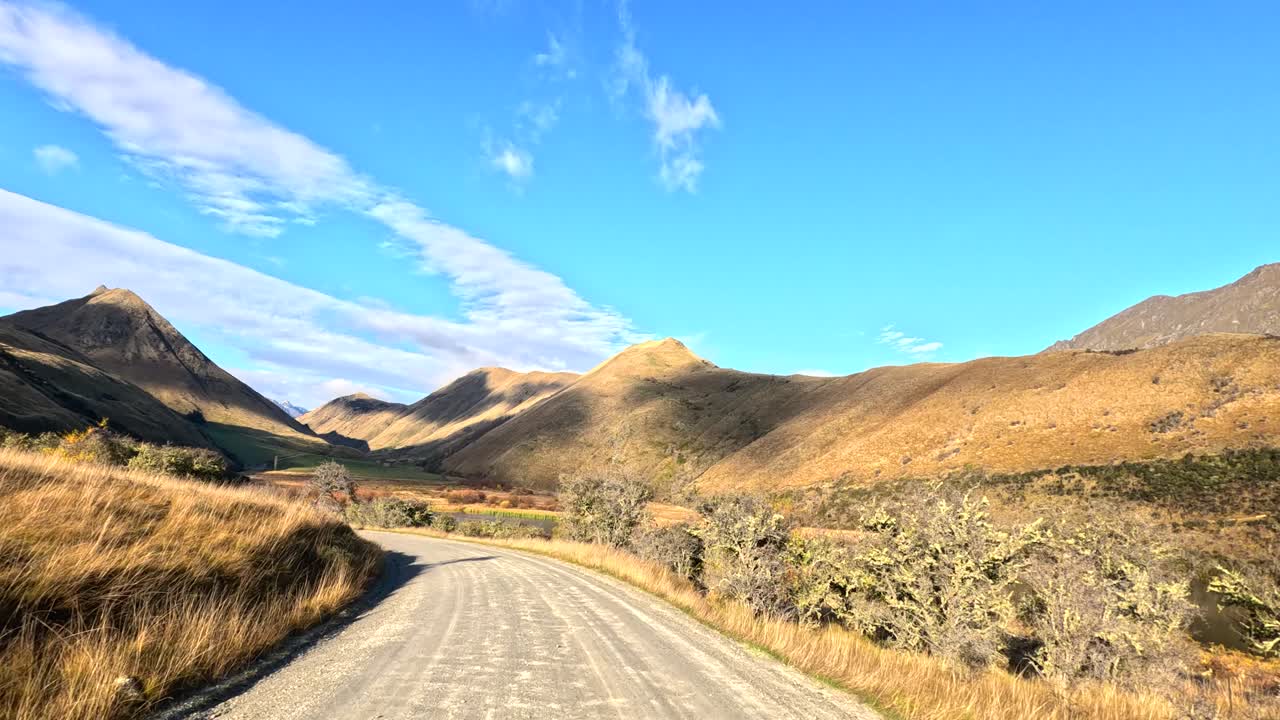 Vehicle travels along gravel road through sunlit mountain landscape, wide angle, steady forward motion