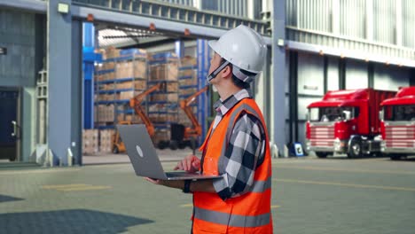 Side View Of Asian Male Engineer With Safety Helmet Working On A Laptop And Looking Around While Standing , Outside of Logistics Distributions Warehouse