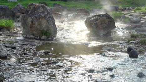 4k slow zoom out shot of of natural hot springs flow along the rocks under sunlight reflection and sun flares, the steam floating up with tree and grass background. lampang,thailand.