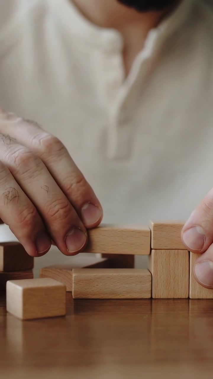 Vertical video: Reaching forward man in henley arranging wooden blocks on tabletop, building steps