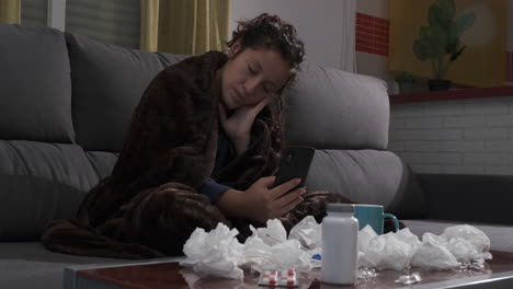 young woman sick on the couch at home, looking for medicine in her mobile phone.