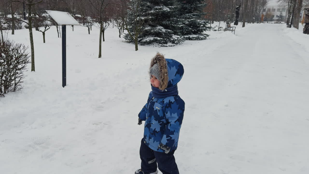 Caucasian baby wearing blue coat with fur hood walks outdoor, speaks and points at something. Walk in the park in winter.