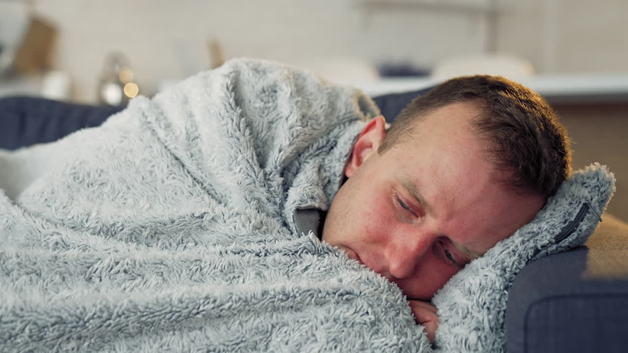 Sick man with a headache during fever. Guy lying in bed covering his head with a warm blanket. Coronavirus pandemic, epidemic. Close-up.