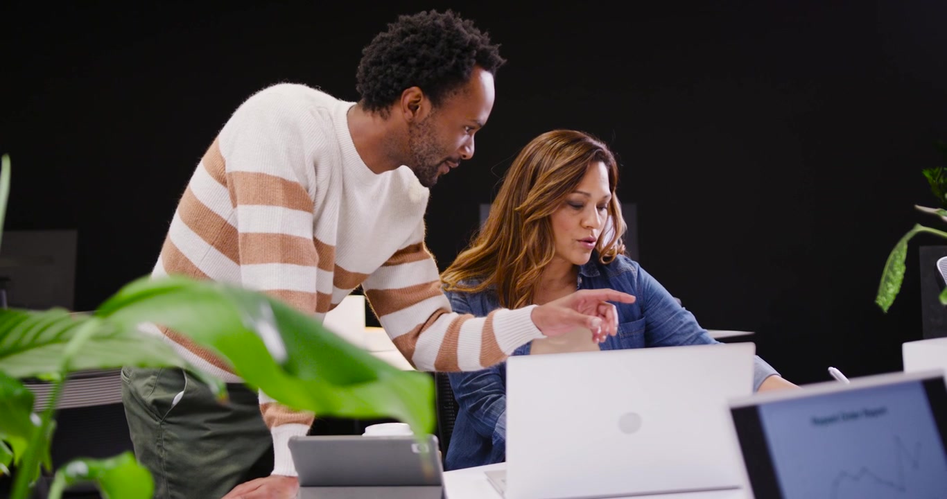 African American coworkers reviewing at office desk, man leaning from left pointing at white laptop