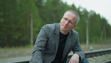 close-up view of a man in a gray jacket and putting on glasses, while seated on a railway track in discomfort, with a blurred background of greenery and tracks