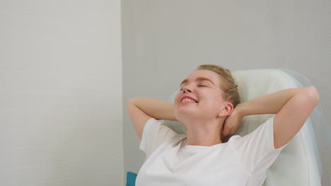 client with hair tied in bun reclines on chair, smiling gently while lifting arms in relaxed motion. scene captures peaceful indoor moment with soft background and natural emotional expression