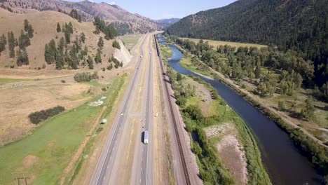 Toma-Aérea-De-Un-Tren-Que-Circula-Junto-A-Un-Río-Y-Una-Carretera-En-La-Costa-Oeste-De-Estados-Unidos-En-Terreno-Montañoso