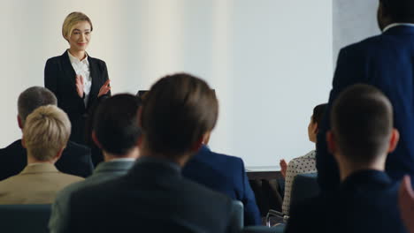 Young-Caucasian-businesswoman-standing-on-a-podium-and-presenting-her-colleague-African-American-man-who-is-sitting-on-a-chair-and-coming-to-the-stage