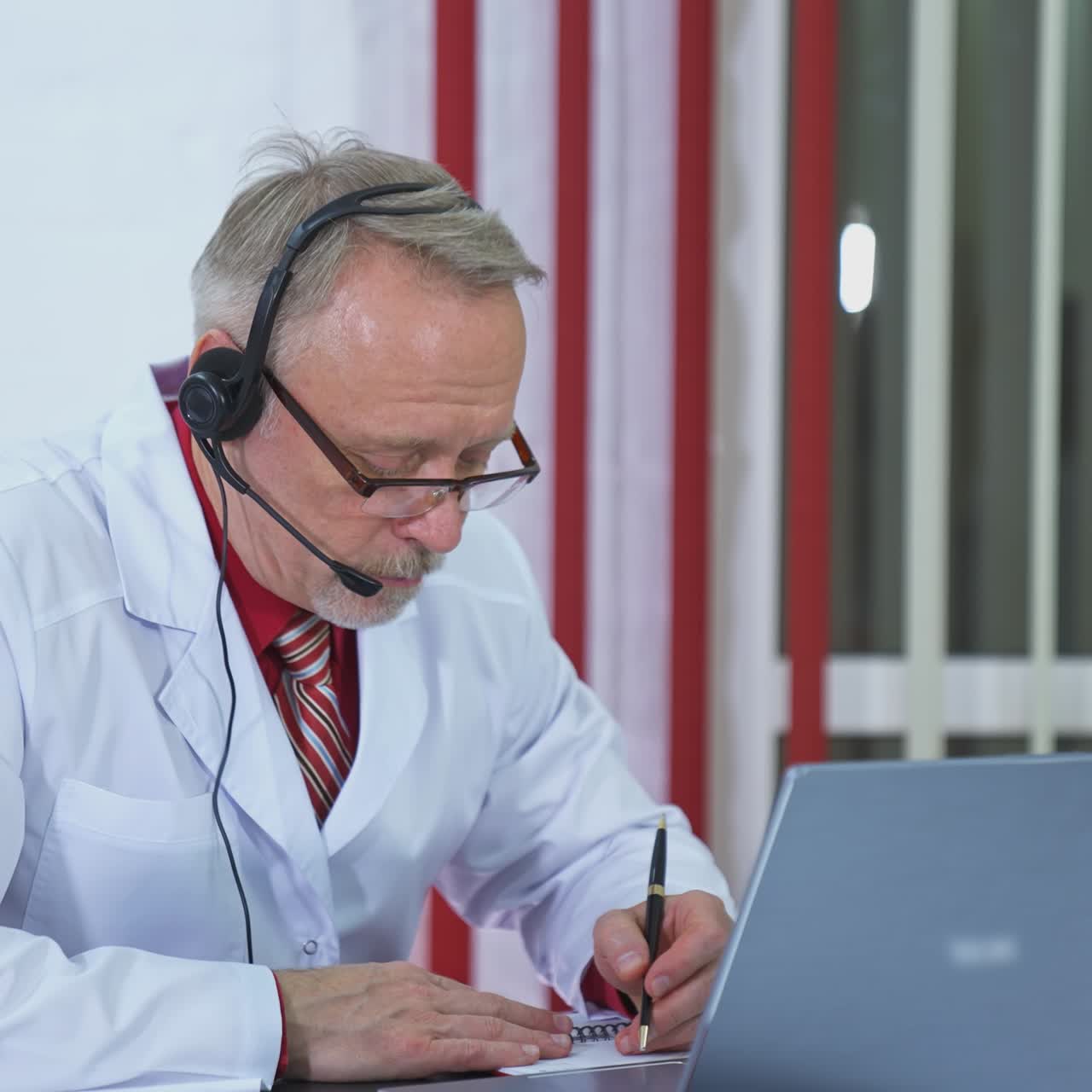 Remote consultation. Senior doctor in medical coat talking online and writing in medical office. Medical specialist consults patient remotely using laptop and headset.