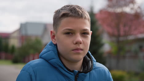 close-up of boy wearing blue jacket outdoors, gazing thoughtfully, with blurred autumn trees and red-roofed house blurred in background