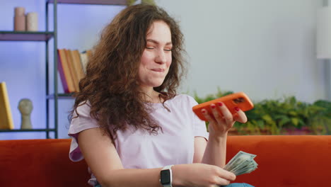 Smiling-happy-young-woman-counting-money-cash-use-smartphone-income-saves-lottery-win-budget-at-home