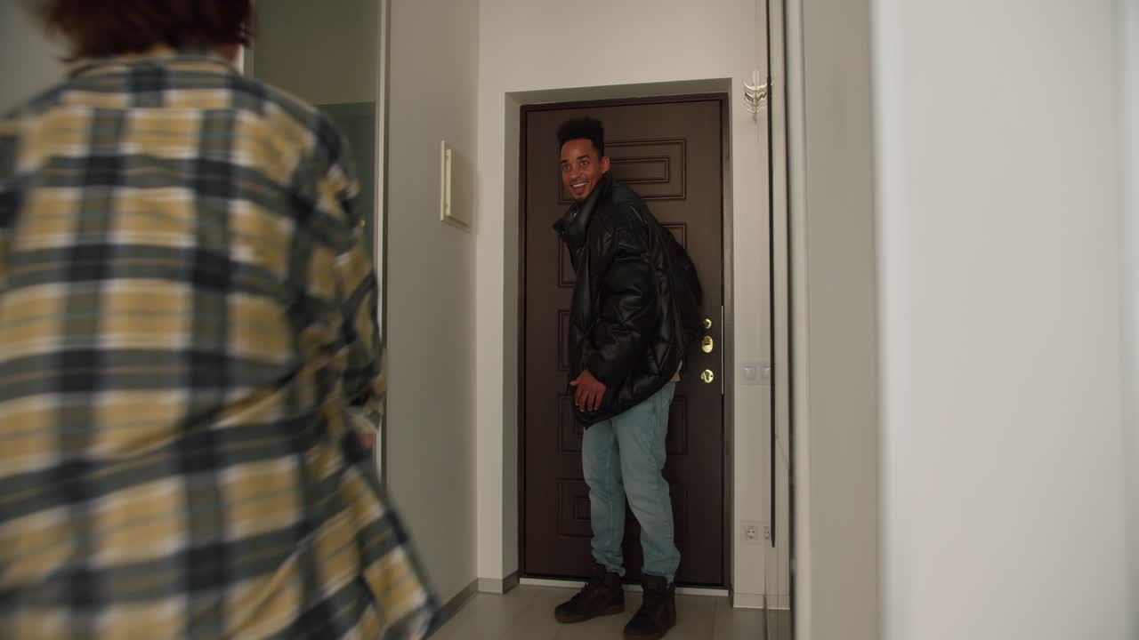 un joven feliz con color de piel negra en una chaqueta negra abre la puerta principal de la casa y llega a casa es recibido por su joven novia adulta morena con un peinado bob en una camisa a cuadros verde y abraza a su novio por la noche