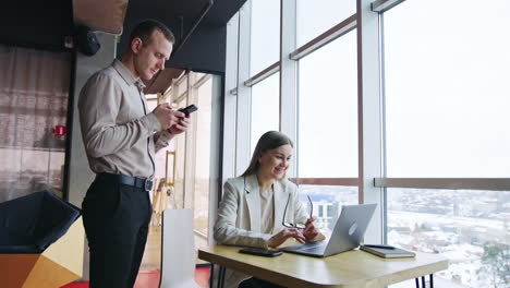 pair of colleagues in office. woman sits at desk in front of laptop addressing man. male workmate looks at the phone in his hands.