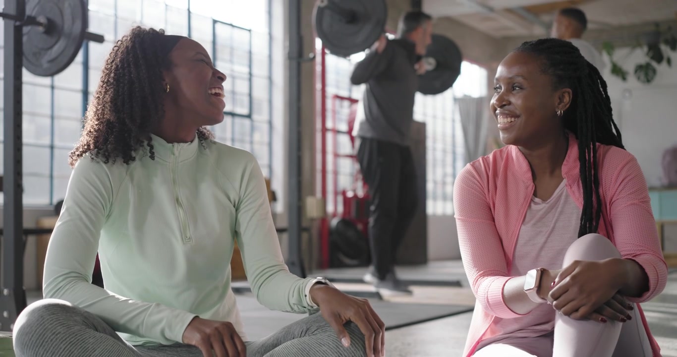 African American women friends chatting laughing on mats in gym between sets mint top barbell