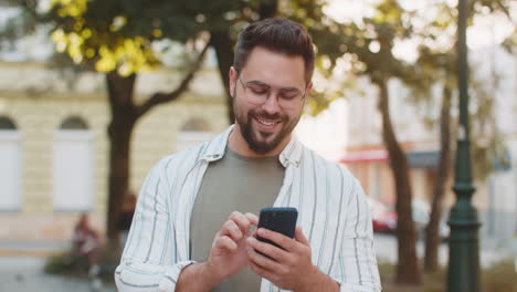 joven sonriente usando su teléfono inteligente escribiendo mensajes de texto desplazando la aplicación de redes sociales en la calle de la ciudad