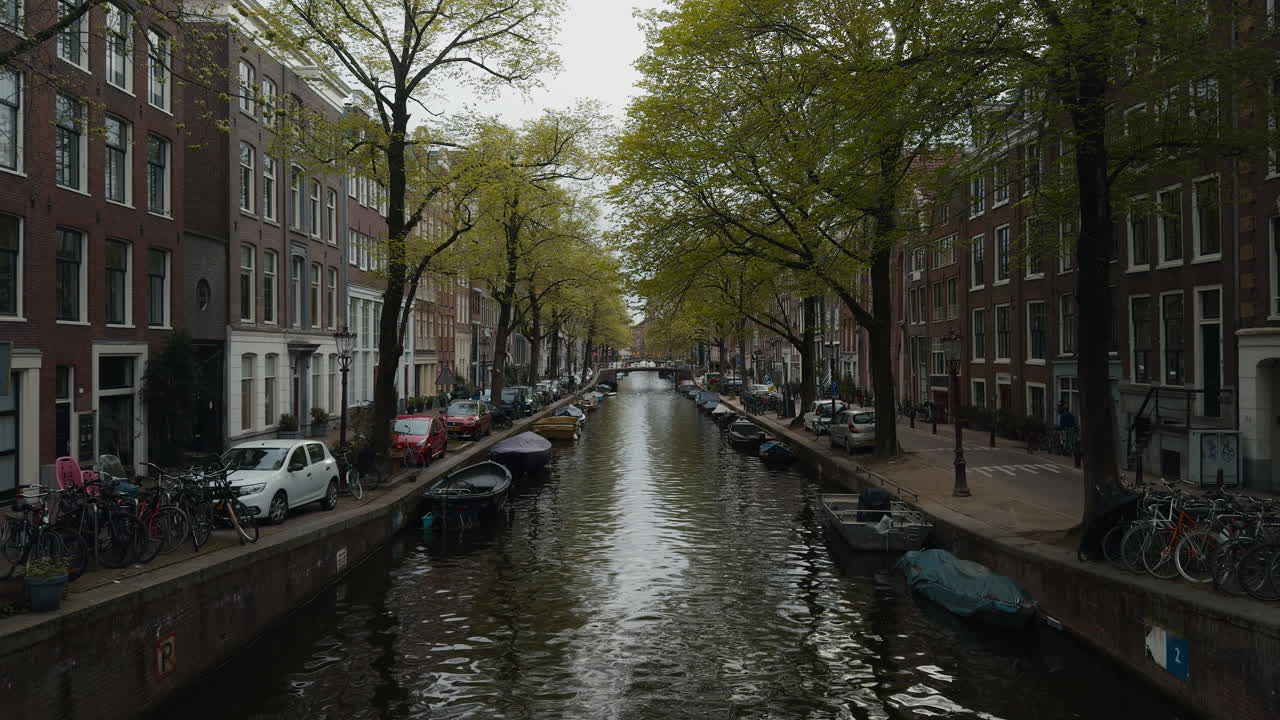 Canal típico de Amsterdã ladeado por casas holandesas históricas, árvores e bicicletas estacionadas, com barcos atracados na água em um dia nublado.