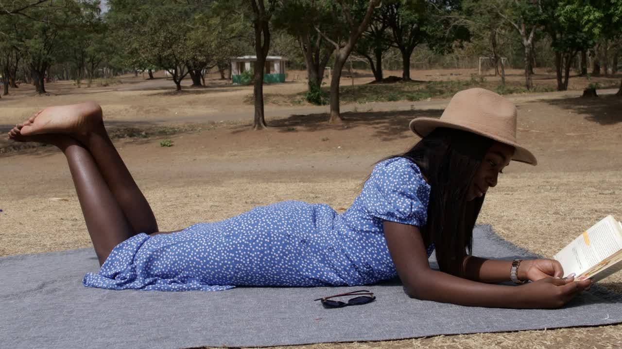 señora en un vestido azul con un sombrero acostado y leyendo un libro en el parque