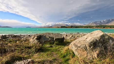 large rocks and thorny plants line a grassy, windswept lake shore with turquoise water, dramatic clouds, and distant snow-capped mountains. subtle camera movement enhances the dynamic, stormy atmosphere