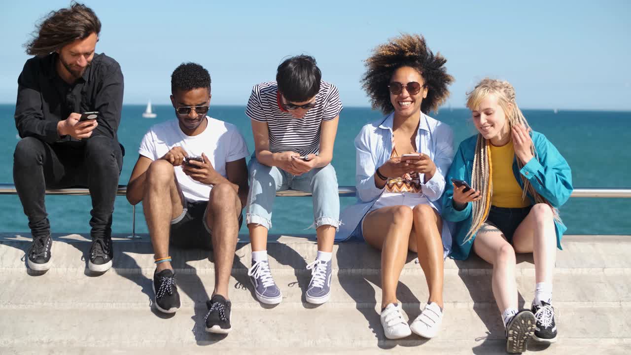 amigos diversos contemporáneos con teléfonos inteligentes en el paseo marítimo