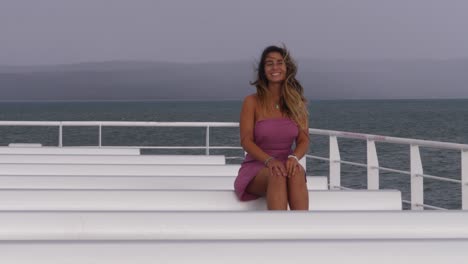 chica vestida sentada en la cubierta sonriendo a la cámara mientras el pelo sopla en el viento - chica en la cubierta del ferry navegando en el océano