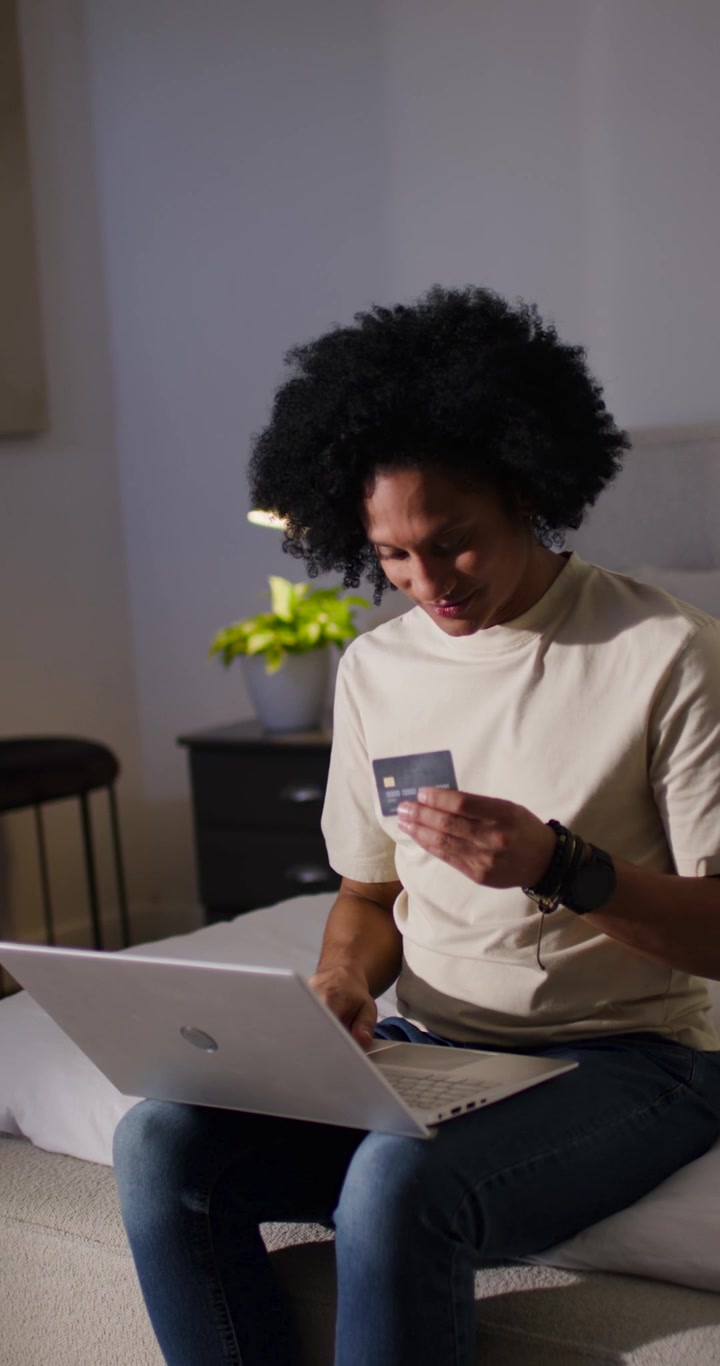 Video verticale: Uomo afroamericano che sorride al laptop, chinato su di esso sul letto e verifica una carta