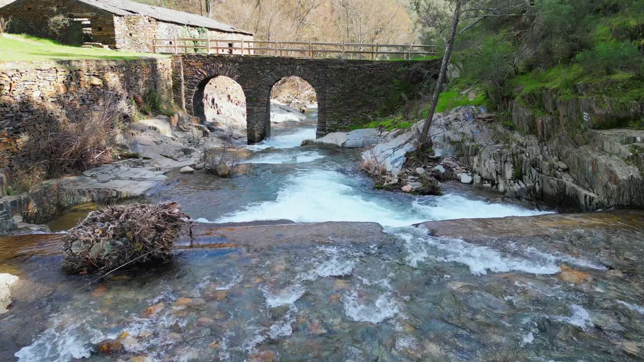 Grabación de dron de un antiguo puente de piedra cruzando un río de montaña cristalino con cañón rocoso y arquitectura tradicional en Extremadura, España. Paisaje de viaje europeo pintoresco y monumento patrimonial