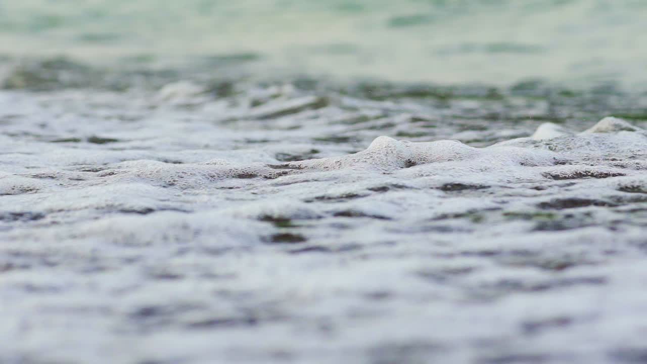 Flowing sea water on the beach with white foam in the evening. The edge of the ocean with foam waves hitting into the shoreline background. Close-up.