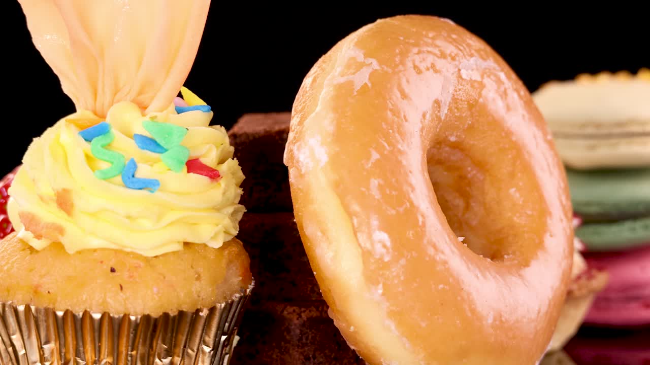 A vibrant selection of baked goods including cupcakes, donuts, macarons, brownies, tarts, and strawberries rotates smoothly against a black background with bright, even lighting